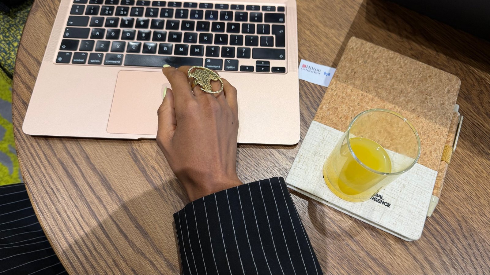 Mantate’s hand, wearing a gold Africa-shaped ring, working on a laptop beside a notebook
